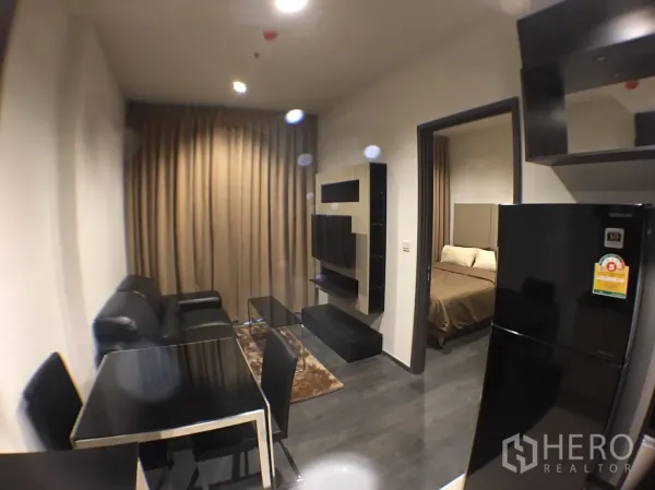 公寓 出租在Watthana, Bangkok - 紧凑客厅，黑色皮沙发、餐桌、电视墙柜，旁边通往卧室并配大冰箱。