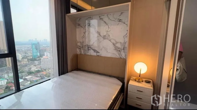 Condominium for rent in Khlong Toei, Bangkok - Cozy bedroom with marble feature wall, bedside lamp, and large city-view window.