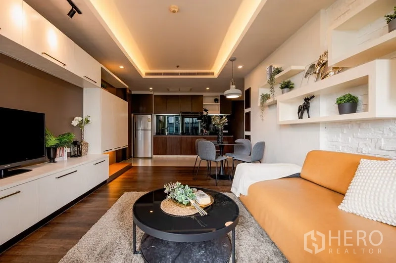 Condominium for sale in Khlong Toei, Bangkok - Open-plan living room with tan sectional sofa, round coffee table, floating shelves, and view to the kitchen-dining under cove lighting.