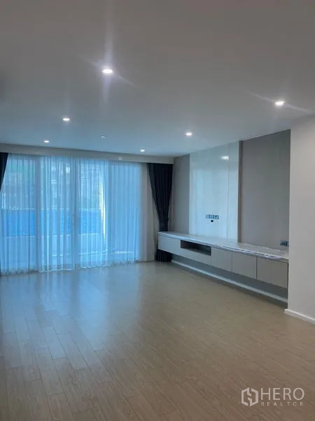 Apartment for rent in Watthana, Bangkok - Bright living room with floor‑to‑ceiling windows, sheer curtains, recessed lights and a built‑in media console on light wood floors.