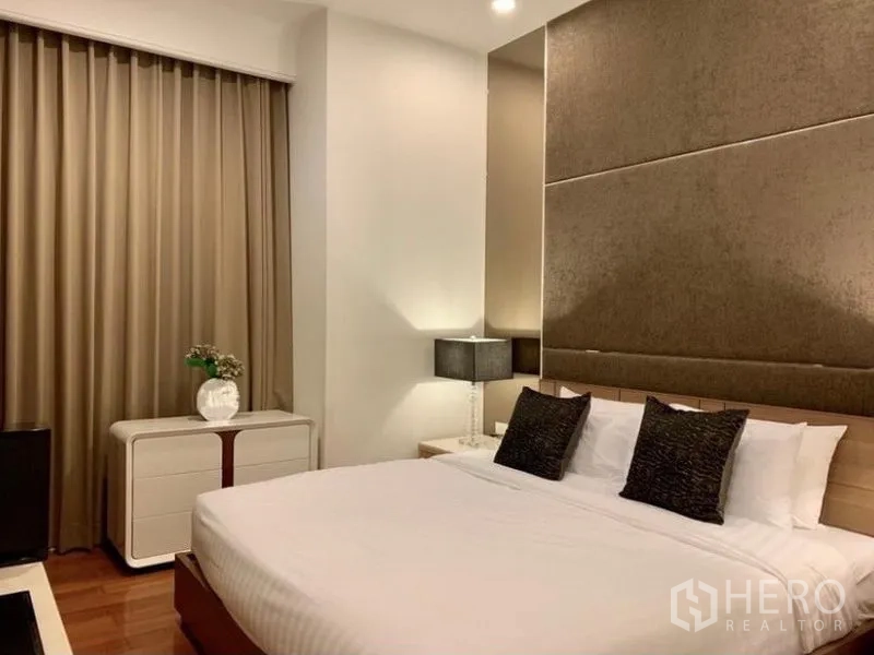 Condominium for rent in Pathum Wan, Bangkok - Primary bedroom with upholstered headboard, king bed with white linens, bedside lamps, dresser, blackout curtains, and wooden flooring.