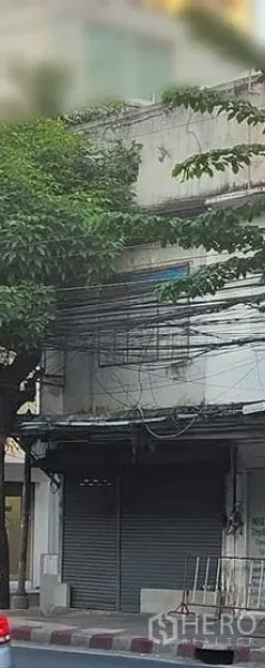 Shophouse for rent in Watthana, Bangkok - Street-front two-storey shophouse exterior with closed metal roller shutter, sidewalk curb, leafy tree and overhead cables in urban Ekkamai, Bangkok.