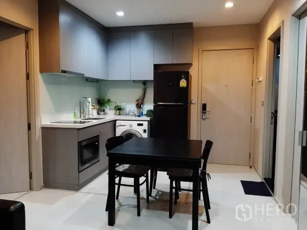 Condominium for rent or sale in Khlong Toei, Bangkok - Modern L-shaped kitchen with upper cabinets, built-in oven, washing machine, fridge and a four-seat dining table.