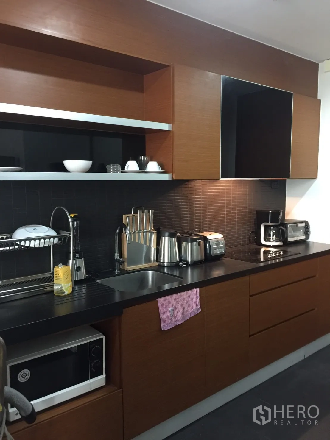 Condominium for rent in Khlong Toei, Bangkok - Modern galley kitchen with black countertop, built-in cabinets, hob and small appliances.