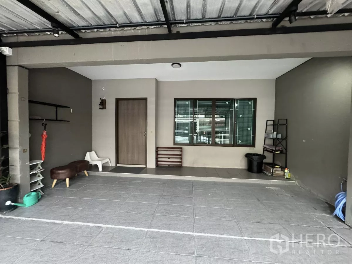 Townhouse for rent in Suan Luang, Bangkok - Covered carport with tiled floor, front door, large window, and shelving along the side wall.
