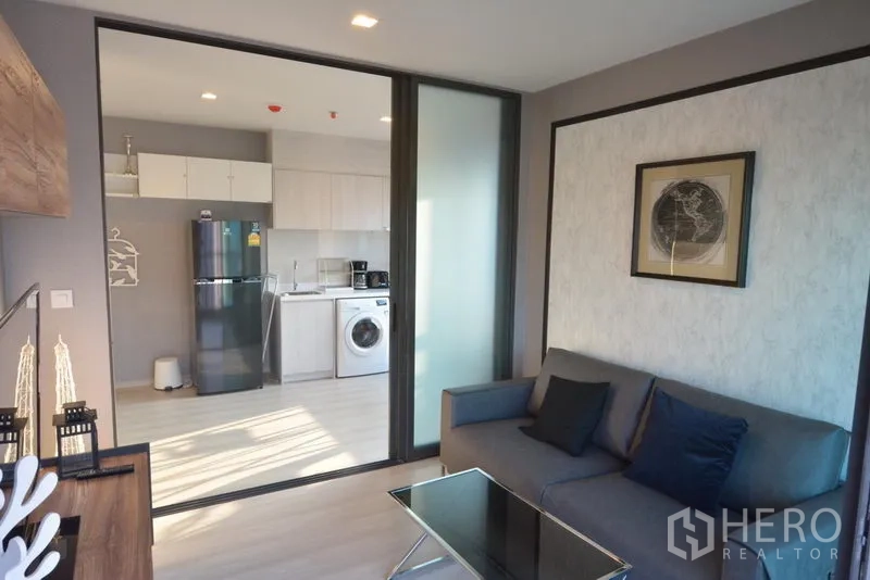 Condominium for sale in Khlong Toei, Bangkok - Cozy living room with gray sofa, glass coffee table, and sliding doors opening to the bright kitchen.