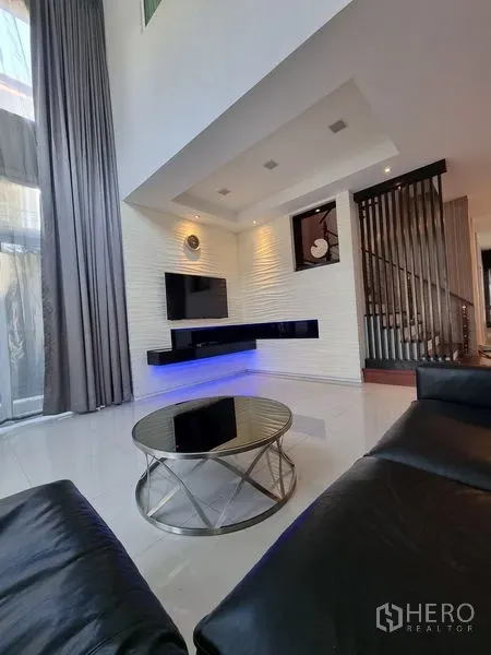 Detached House for sale in Chatuchak, Bangkok - Double-height living room with wall-mounted TV, LED-lit shelves and a round glass coffee table.
