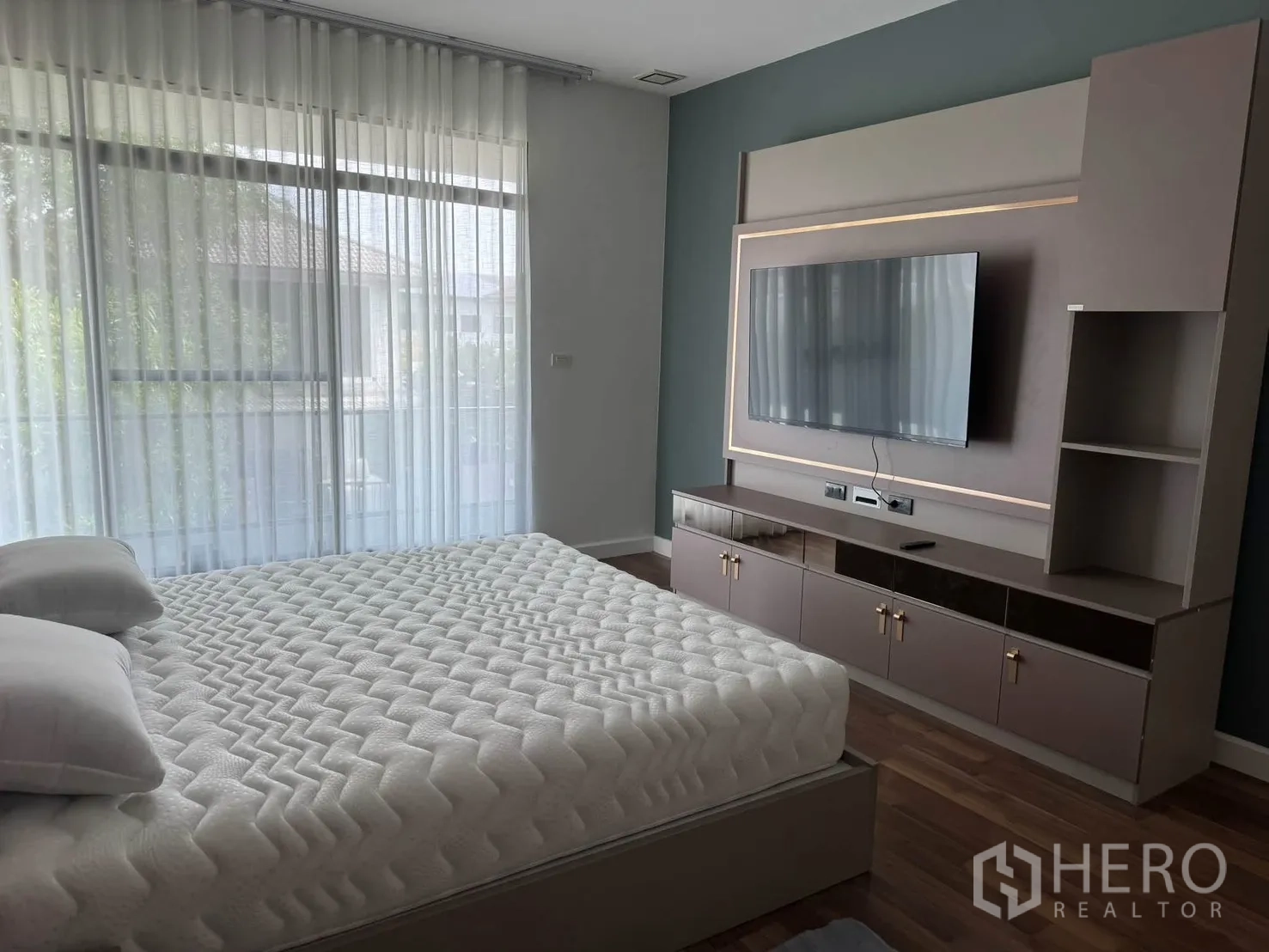 Detached House for rent in Prawet, Bangkok - Master bedroom wall with mounted TV, storage cabinetry, and sheer curtains to a balcony.
