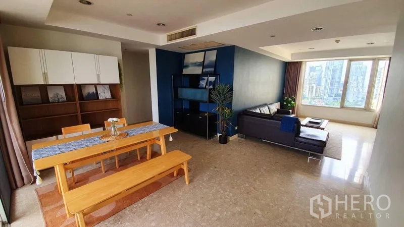 Condominium for rent in Watthana, Bangkok - Open‑plan living and dining room with wooden table, blue accent wall and large city‑view windows.