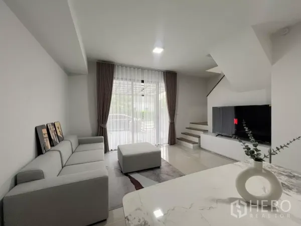 Townhouse for rent in Bang Phli, Samut Prakan - Open-plan lounge with sofa, ottoman, TV unit and staircase facing the front patio.