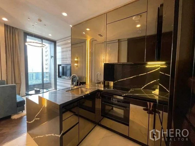 Condominium for rent in Khlong Toei, Bangkok - Sleek U‑shaped kitchenette with black stone countertops, built‑in oven and mirrored cabinets opening to the living area.