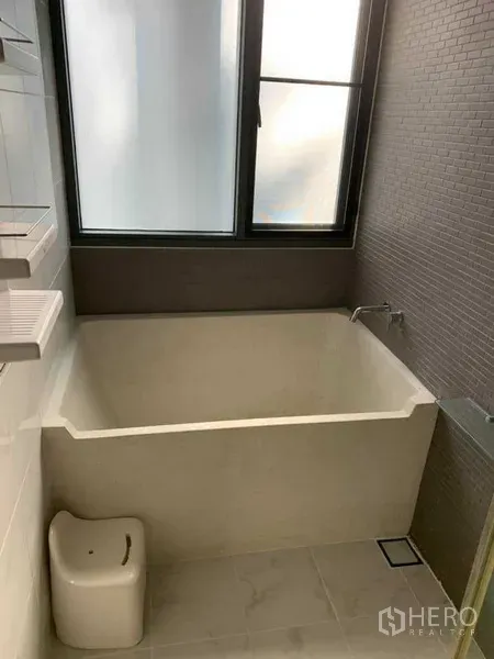 公寓 出租在Watthana, Bangkok - 现代浴室，深浸浴缸与磨砂窗，灰色瓷砖墙面。