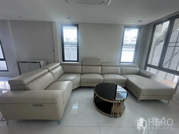 Townhouse for rent in Watthana, Bangkok - Bright living room with large L-shaped leather sofa and round coffee table beside tall windows.
