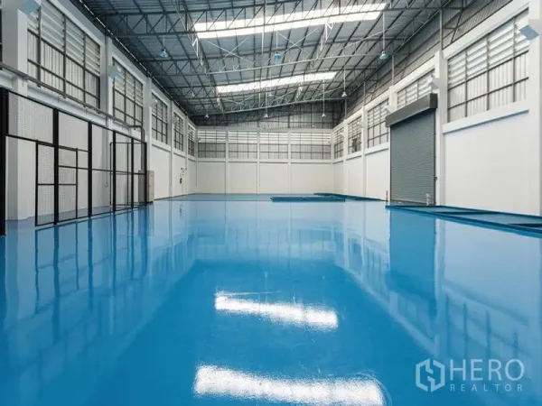 Factory for rent in Bang Bo, Samut Prakan - High‑ceiling warehouse interior featuring glossy blue epoxy floor, skylights, ventilation panels and an oversized roller shutter.