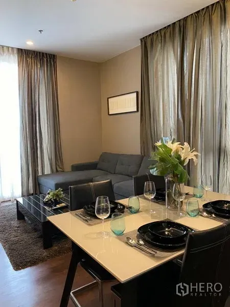 Condominium for rent in Watthana, Bangkok - Dining area for four beside a gray sectional sofa, coffee table and tall sheer curtains allowing natural light.
