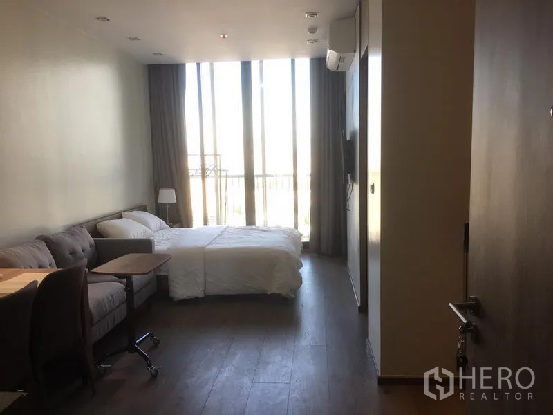 公寓 出租在Khlong Toei, Bangkok - 明亮房间，落地窗与阳台栏杆，配床、沙发和壁挂电视。
