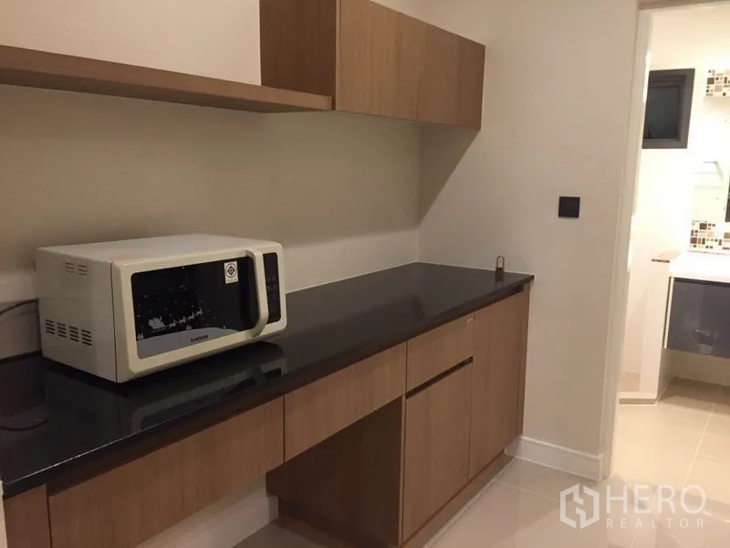 Detached House for rent in Bang Kapi, Bangkok - Pantry with long black countertop, wood cabinets and microwave, leading toward a bathroom.
