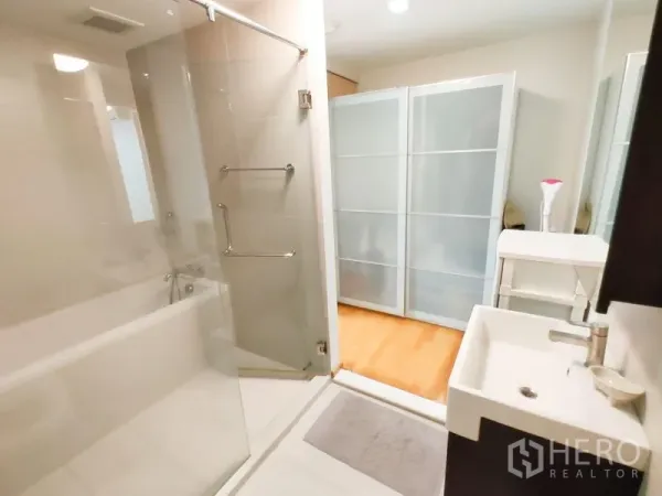 Condominium for rent in Khlong Toei, Bangkok - Glass-enclosed shower and bathtub beside a vanity, opening toward a room with frosted sliding doors.