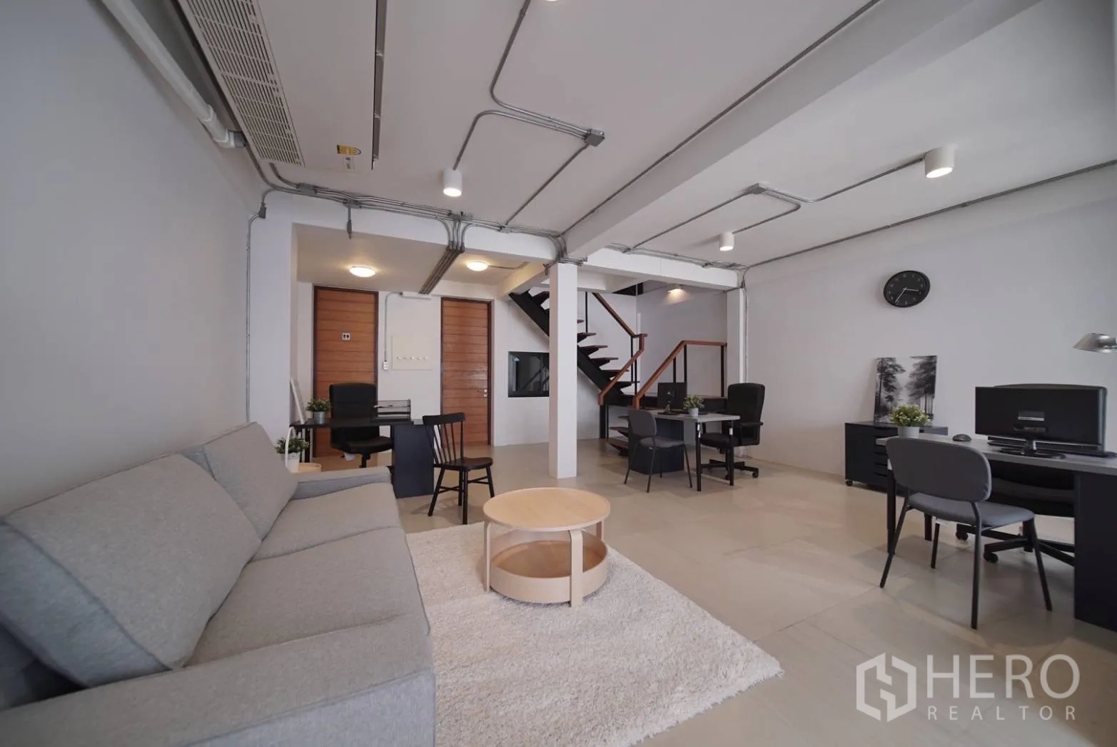 Townhouse for rent in Watthana, Bangkok - Open-plan ground floor with sofa, multiple desks, exposed ceiling conduits and staircase.