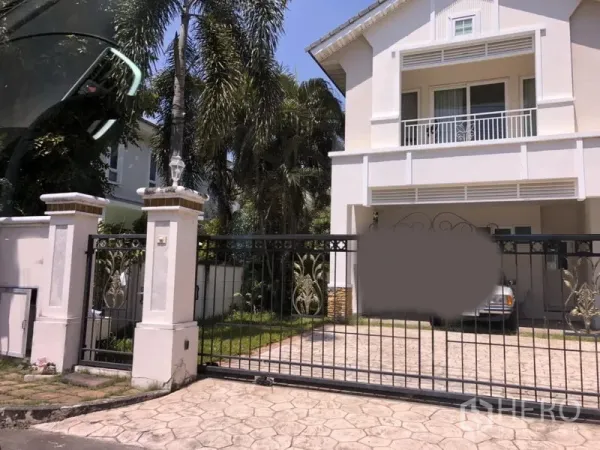 Detached House for  in Lat Krabang, Bangkok - Two-storey white detached house in Lat Krabang, Bangkok with balcony, secure metal gate, paved driveway, covered carport and palm-lined lawn.
