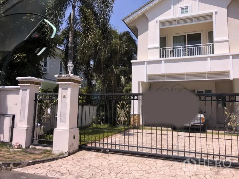 Detached House for  in Lat Krabang, Bangkok - Two-storey white detached house in Lat Krabang, Bangkok with balcony, secure metal gate, paved driveway, covered carport and palm-lined lawn.