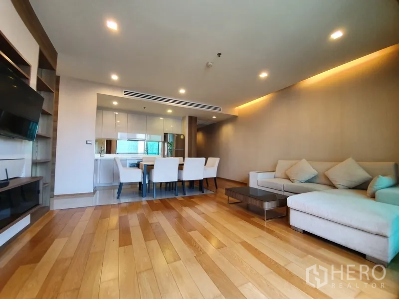 Condominium for rent in Bang Rak, Bangkok - Spacious living area with wood floors, L-shaped sofa, and dining zone beside a sleek white kitchen.
