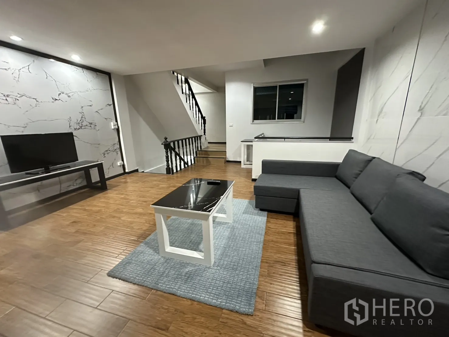 Home Office for rent in Khlong Toei, Bangkok - Open living area with gray sectional sofa, TV stand, and wood floors.