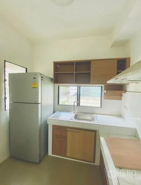 Detached House for rent or sale - Compact closed kitchen with L‑shaped counter, sink, stainless refrigerator, windows and overhead cabinets.