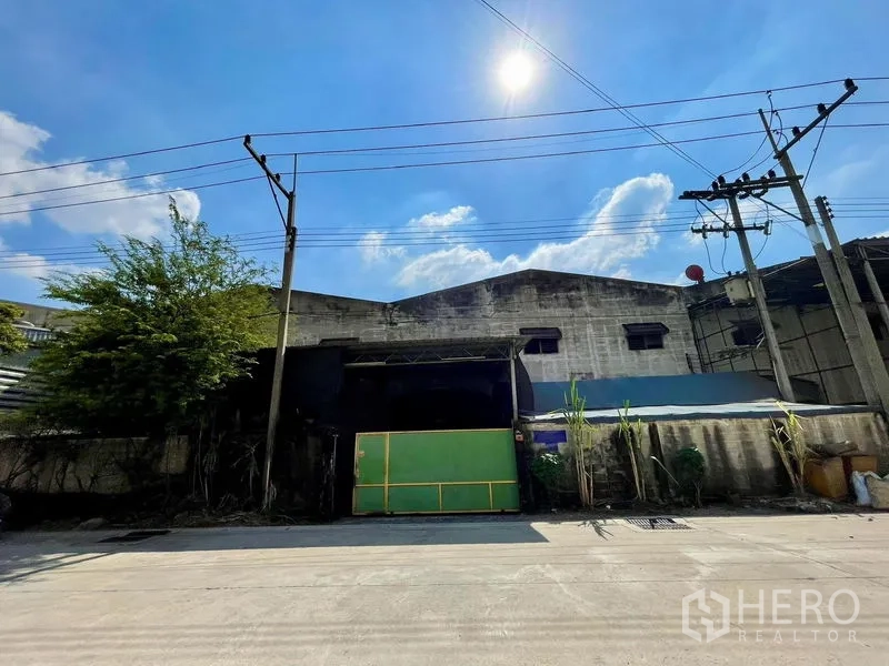 คลังสินค้า ให้เช่าในKrathum Baen, Samut Sakhon - ด้านหน้าอาคารโกดัง ประตูเลื่อนสีเขียว กำแพงรั้ว และสายไฟเหนือถนน