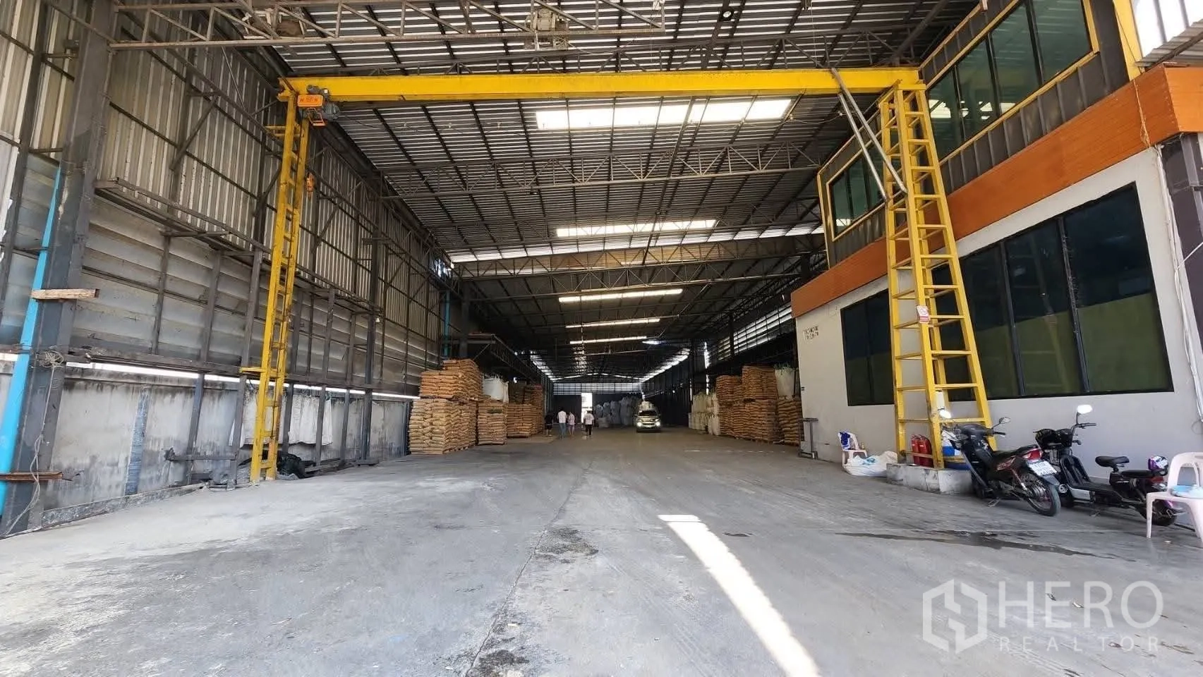 Warehouse for rent in Bang Phli, Samut Prakan - Long central aisle of the warehouse with gantry beam, stacked materials, and a vehicle under the lit steel roof.