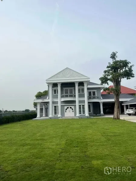 บ้าน ขายในBang Na, Bangkok - ด้านหน้าบ้านเดี่ยวสไตล์นีโอคลาสสิกเวลากลางวัน มีเสาสี่ต้นและสนามหญ้าใหญ่