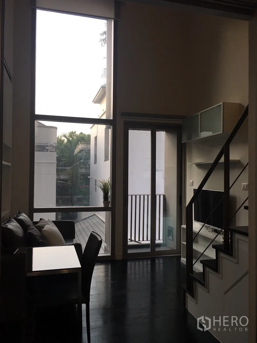 Condominium for rent in Khlong Toei, Bangkok - Double-height living room with floor-to-ceiling windows, balcony door, staircase and TV cabinet.