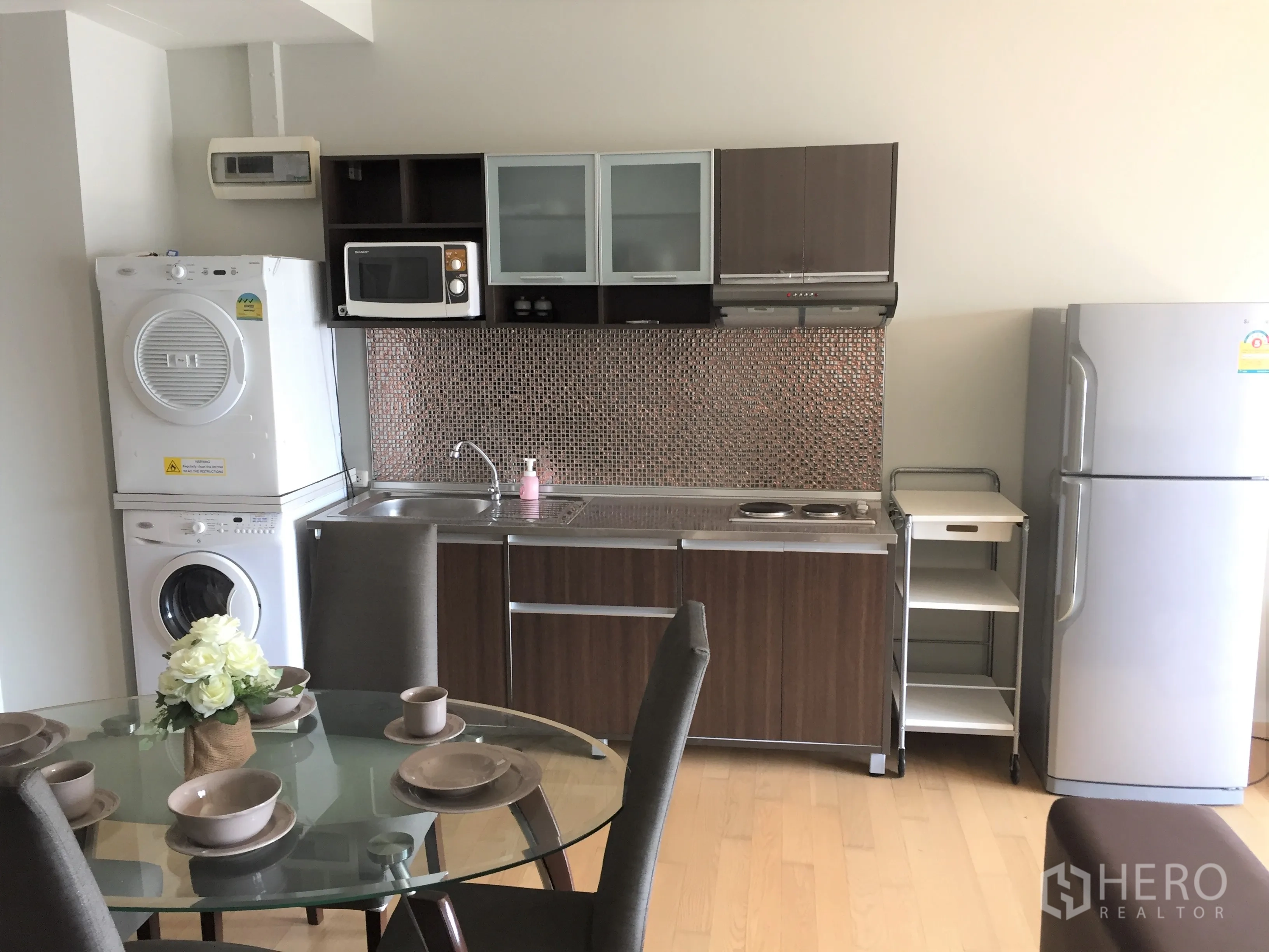 Condominium for rent in Watthana, Bangkok - Compact condo kitchen with mosaic backsplash, electric cooktop, stacked washer‑dryer, microwave, fridge and a glass dining table.