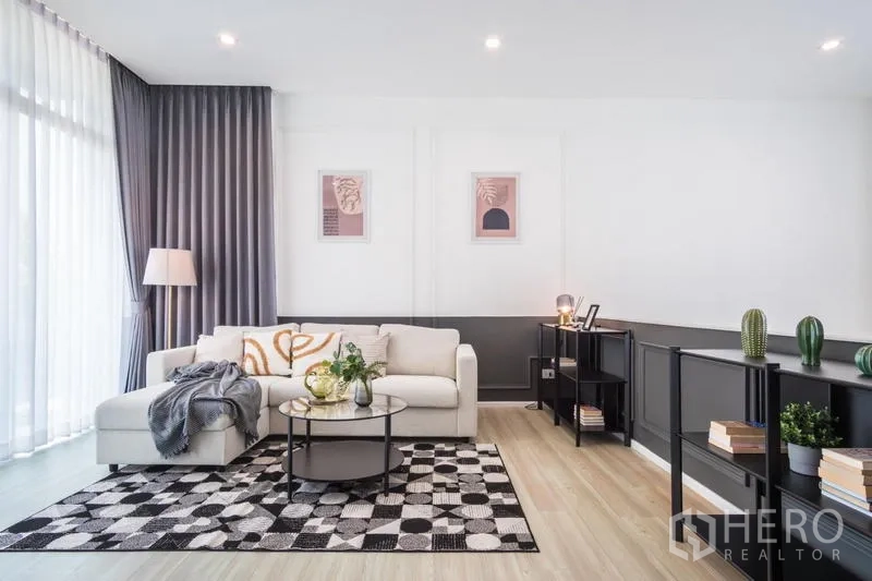 Townhouse for rent or sale in Suan Luang, Bangkok - Living room with L-shaped sofa, round coffee table, patterned rug, and open shelving.