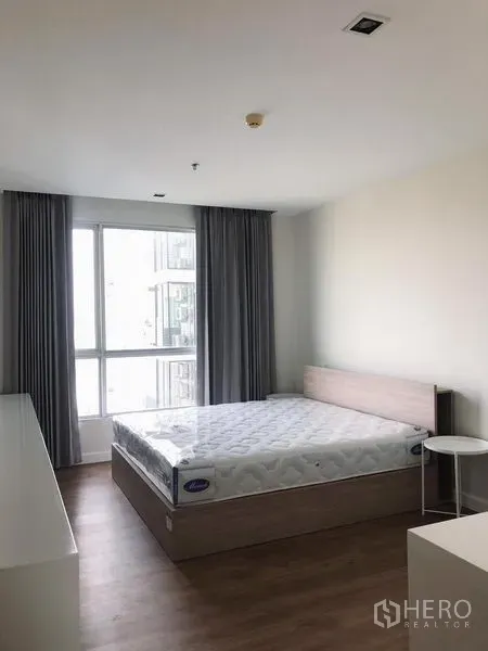 Condominium for rent in Bang Rak, Bangkok - Spacious bedroom with large window, grey curtains and light-wood bed frame with mattress.