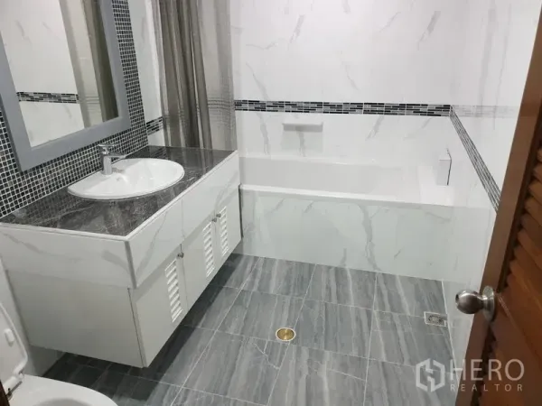Condominium for rent in Watthana, Bangkok - Modern bathroom with marble-style tiles, a vanity sink, and a bathtub with shower curtain.