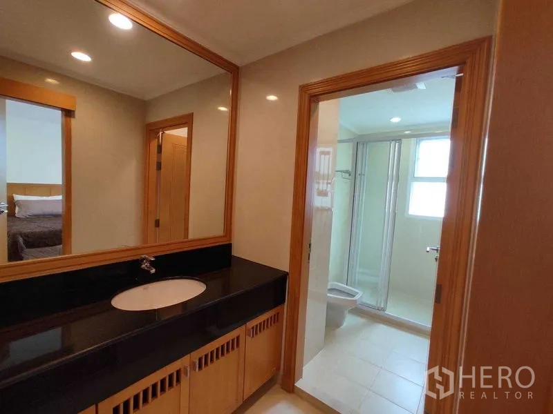 Condominium for rent in Khlong Toei, Bangkok - Vanity with black countertop and large mirror leading to a bathroom with glass shower enclosure.