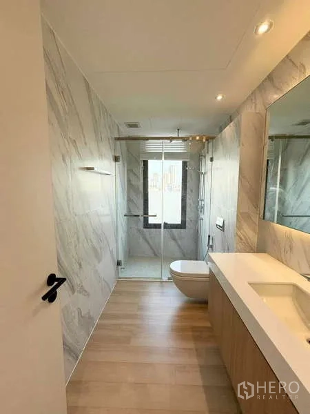 Condominium for rent in Watthana, Bangkok - Modern bathroom with marble walls, long vanity, glass-enclosed rain shower, and city-view window.