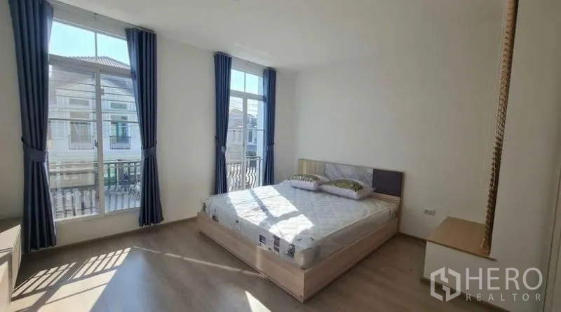 Townhouse for rent in Prawet, Bangkok - Sunlit master bedroom with large windows, blue curtains and a small balcony view.