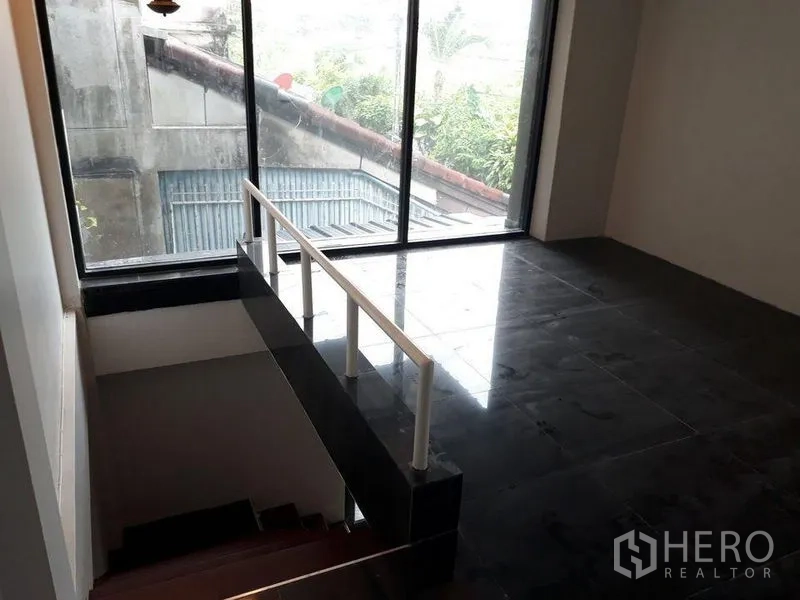 Townhouse for sale in Bang Kapi, Bangkok - Upper landing with dark tile flooring and a full-height window overlooking neighboring roofs.