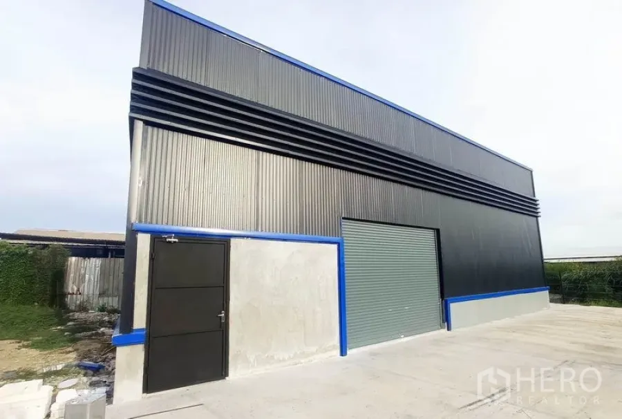 Warehouse for rent in Saphan Sung, Bangkok - Modern corrugated-metal warehouse exterior with blue trim, roller shutter and side door on a concrete yard in Bangkok.