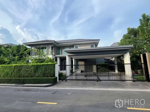บ้าน ให้เช่าหรือขายในLat Krabang, Bangkok - หน้าบ้านมีกำแพงรั้วเหล็ก โรงจอดรถมีหลังคา และพุ่มไม้ตัดแต่งริมถนน