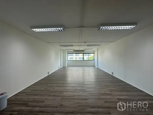 Office Space for rent in Bangkok - Empty office space with ceiling lights, white walls and a windowed facade overlooking nearby buildings.