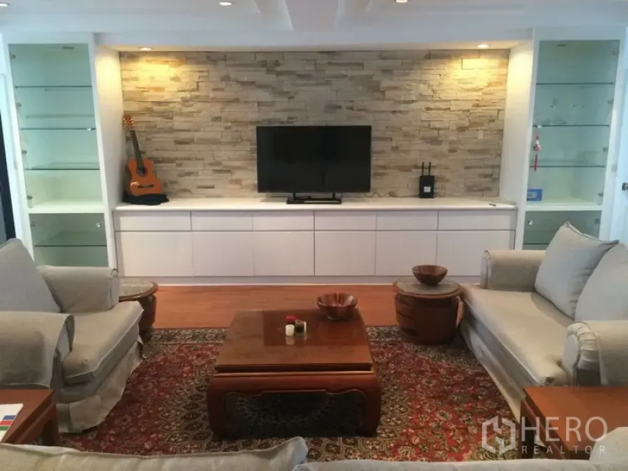 Condominium for rent in Khlong Toei, Bangkok - Living room focal wall with stacked-stone cladding, TV, glass display shelves and two sofas around a rug.