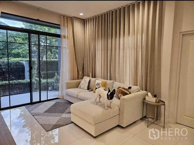 Detached House for sale in Bang Kruai, Nonthaburi - Bright living room with L-shaped sofa, floor-to-ceiling curtains and sliding doors to the patio.