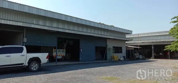 Warehouse for rent in Khlong Sam Wa, Bangkok - Metal-clad warehouse bays with roller shutters, covered loading area and gravel yard with pickup truck in front.