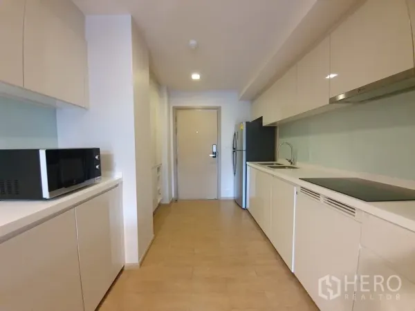 公寓 出租在Watthana, Bangkok - 修长厨房，白色柜体、玻璃防溅板、电磁炉、微波炉与大冰箱。