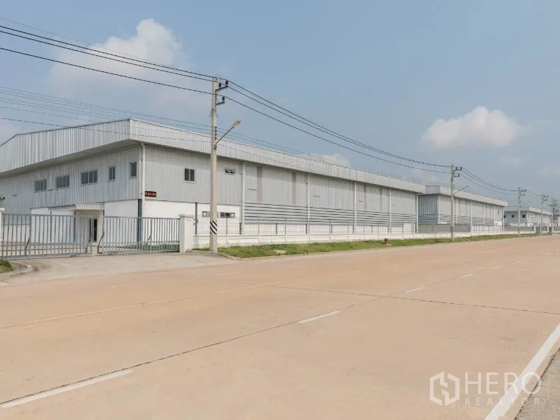 工厂 出租在Si Racha, Chon Buri - 宽阔工业道路旁的浅灰色金属厂房外观，带围栏与电线杆