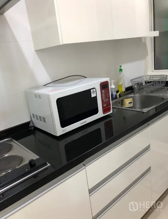Condominium for rent or sale in Khlong Toei, Bangkok - Compact white kitchen with black countertop, sink, microwave and electric hob.