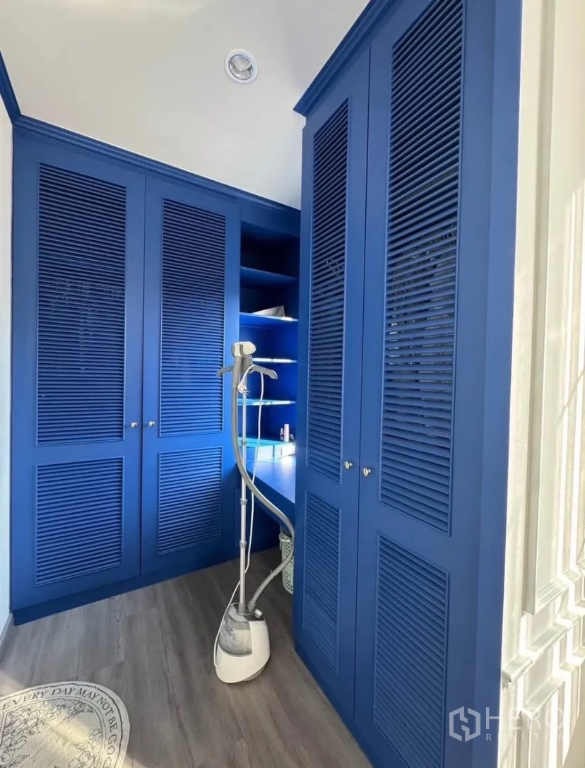 Detached House for rent or sale in Lat Krabang, Bangkok - Walk-in closet featuring bold blue built-in wardrobes, open shelving, and a garment steamer on wood floors.